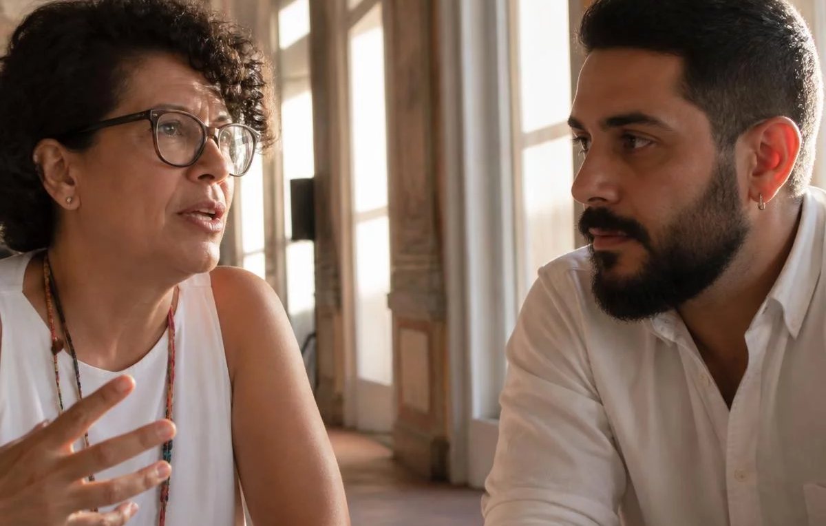 Duas pessoas sentadas de frente uma para a outra à mesa: uma fala, a outra registra por escrito
